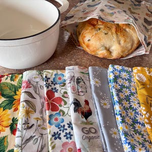 May include: A white enameled cast iron pot, a loaf of bread in a chicken-patterned fabric bag, and seven folded fabric napkins featuring various floral, rooster, and organic-themed prints in yellow, blue, red, white, and gray colors.