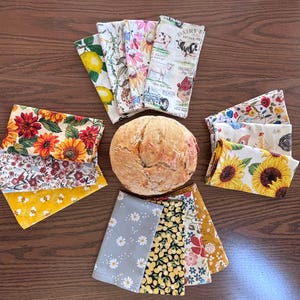 May include: A round loaf of bread surrounded by a collection of colorful floral and farm-themed cloth napkins. The napkins feature various patterns, including sunflowers, lemons, and daisies. The arrangement is set on a dark wooden surface.