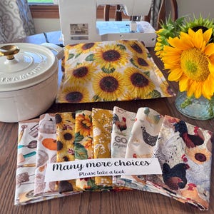 May include: A white enameled cast iron Dutch oven, a sewing machine, a glass vase with yellow sunflowers, and a stack of folded fabric squares featuring patterns of sunflowers, pumpkins, and chickens with a sign reading Many more choices Please take a look.