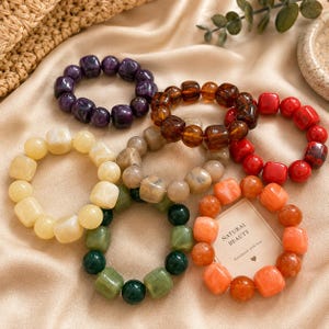 May include: Six circular bracelets made of polished, chunky, square-shaped beads in purple, amber, red, cream, olive green, and orange. A small rectangular card with the text "NATURAL BEAUTY Handmade with love" is tucked inside the orange bracelet.