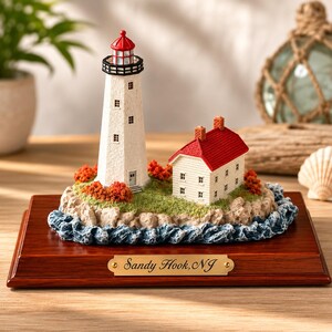 May include: A miniature Sandy Hook, NJ lighthouse figurine featuring a white tower with a red top, a white house with a red roof, green landscaping, and rocky terrain surrounded by blue waves, all mounted on a rectangular wooden base.