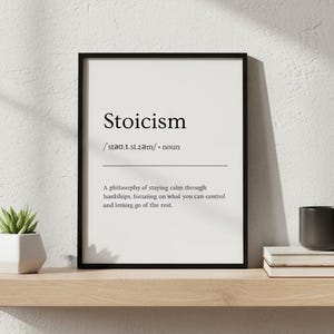 以下が含まれることがあります： 「ストイシズム」の定義が黒字で書かれた額入りのプリント。明るい色の木製の棚に、小さな植物、黒いマグカップ、2冊の本と一緒に置かれています。壁は白です。