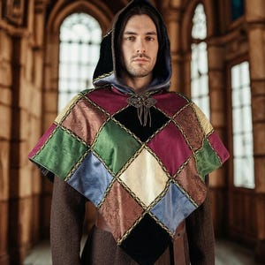 Op de afbeelding: Een cape met capuchon voorzien van een ruitvormig patchworkpatroon in de kleuren bordeauxrood, groen, blauw, zwart en goud. Het kledingstuk wordt bij de hals vastgemaakt met een decoratieve metalen sluiting en gedragen over een bruine tuniek.