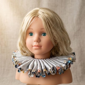 Op de afbeelding: Een pop met blond haar en blauwe ogen draagt een lichtgrijze rucheskraag. De kraag heeft een geplooid ontwerp met een donkere kralenrand aan de binnenkant en een kleurrijk bloemenpatroon zichtbaar langs de buitenste randen van de stof.