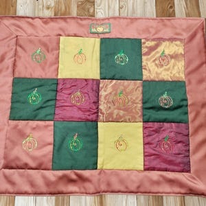 Op de afbeelding: Een gewatteerde mat met een patchwork ontwerp met geborduurde pompoenen. De mat heeft een stoffige roze rand en vierkanten van groene, gele en bordeauxrode stof. De pompoenen zijn geborduurd in tinten oranje en groen.
