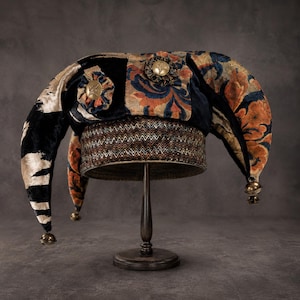 Op de afbeelding: Een narrenmuts met meerdere punten van zwart, oranje en blauw fluweel met een bloemenpatroon. De muts heeft een brede, gestructureerde band met een chevronpatroon, twee ronde messingkleurige sierspelden en kleine metalen belletjes aan de punten.