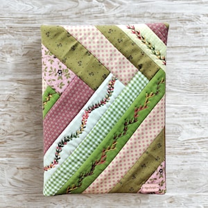 Op de afbeelding: Een gewatteerde boekomslag met een patchwork-ontwerp. Het heeft diagonale strepen in verschillende patronen, waaronder bloemen, stippen en ruitjes, in roze, groen en wit. Geborduurde details voegen een decoratief tintje toe.