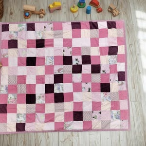 Op de afbeelding: Een handgemaakte patchworkquilt in roze en bordeaux tinten, gerangschikt in een rasterpatroon. De quilt heeft verschillende vierkante stoffen met bloemen en effen stoffen. De quilt is omgeven door houten speelgoed, waaronder een speelgoedauto en een hond.