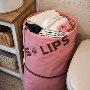 May include: A cylindrical pink fabric laundry hamper with a drawstring closure and white rope cord. The exterior features the dark brown text "S LIPS" with a small geometric symbol between the letters. The hamper is filled with folded clothing.