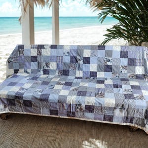 Op de afbeelding: Een patchwork quilt in tinten blauw, grijs en wit bedekt een bank. De quilt heeft verschillende vierkante patronen, waaronder bloemen-, ruit- en stippelontwerpen. De bank, op wielen, staat buiten in de buurt van een strand.