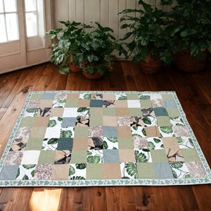 Puede incluir: Un edredón patchwork rectangular con un diseño de hojas tropicales en tonos verdes, beige y blanco. El edredón tiene un borde estampado y está colocado sobre un suelo de madera. Dimensiones aproximadas: 150 cm x 200 cm.