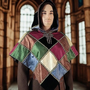 May include: A multi-colored patchwork cape with a hood, featuring squares of green, blue, burgundy, and brown fabrics. The cape is trimmed with gold-colored braid and has a decorative clasp at the neckline. The mannequin is wearing a brown outfit.