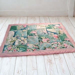 May include: A rectangular patchwork quilt with a floral pattern in shades of green, blue, and pink. The quilt features a pink border and is placed on a light-colored wooden surface. The floral design includes roses and other flowers.