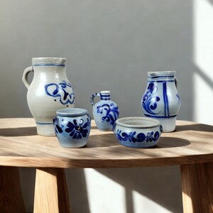 Op de afbeelding: Een verzameling keramisch aardewerk met blauwe bloemmotieven op een lichtbeige achtergrond. De set bevat een hoge kan, een kleine vaas, een kruik met handvat, een kleine kom en een kleine pot. Het aardewerk staat op een ronde houten tafel.