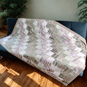 Op de afbeelding: Een patchwork quilt met verschillende patronen in grijstinten, beige en lavendel, gedrapeerd over een donkerblauwe bank. De quilt heeft een lichtbeige rand. De kamer heeft een parketvloer en potplanten.