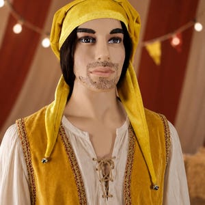 May include: A yellow velvet jester hat with long, pointed ends and a matching vest. The vest has gold trim and is worn over a cream-colored shirt with a laced neckline. The background includes a circus tent and string lights.