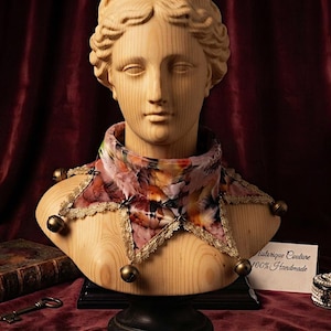May include: A light wooden bust of a classical figure wearing a multi-colored floral print neck collar with gold lace trim and small brass bells attached to the points. A white card with the text "Historique Couture 100% Handmade" is visible.