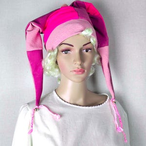Könnte beinhalten: Ein farbenfroher Narrenhut in Rosa- und Lilatönen. Der Hut hat lange, spitze Spitzen und ist mit rosa Quasten und kleinen goldenen Perlen verziert. Der Hut ist aus weichem Stoff gefertigt und hat ein verspieltes, skurriles Design.