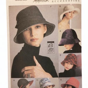 Op de afbeelding: Vogue Accessoires patroon 7460 met verschillende hoedontwerpen. De afbeelding toont een vrouw die een donkergrijze bucket hat draagt, met andere hoedstijlen op de achtergrond. Het patroon bevat alle maten.