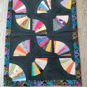 Op de afbeelding: Een handgemaakte quilt met een waaierpatroon. De quilt heeft een zwarte achtergrond met kleurrijke waaier-vormige ontwerpen in verschillende kleuren. De randen zijn afgewerkt met een stof met bloemenpatroon. De waaier vormen zijn omrand met turquoise stiksels.