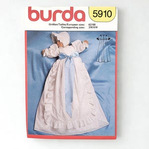 May include: A Burda pattern envelope featuring a baby's christening gown. The gown is white with a light blue ribbon and a matching bonnet. The envelope has the number 5910 and size information.