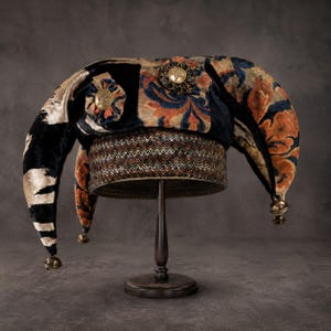 May include: A multi-pointed hat featuring a mix of black velvet and orange and blue floral patterned fabric. The base has a metallic chevron-patterned band. Two gold-toned circular ornaments are attached to the front, and small bells hang from the tips.