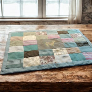 Op de afbeelding: Een patchwork quilt met een verscheidenheid aan stoffen in tinten blauw, groen, bruin en crème. De quilt heeft een gestikt paardenontwerp in het midden. De quilt is rechthoekig en heeft een blauwe rand.