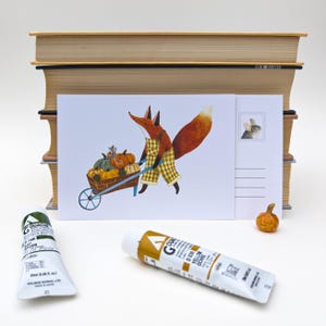 Può includere: Una cartolina stravagante con una volpe con un grembiule a scacchi che spinge una carriola piena di zucche. La cartolina è ambientata contro una pila di libri e materiali artistici, inclusi tubi di vernice. È presente anche una piccola statuina di zucca.
