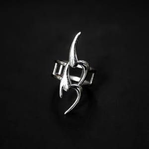Puede incluir: Un anillo plateado con un diseño abstracto único. El anillo presenta una serie de elementos curvos y puntiagudos que crean una estética moderna y llamativa. El anillo se encuentra sobre un fondo oscuro, resaltando su brillo metálico.