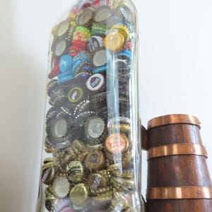 May include: A clear glass bottle filled with a variety of colourful bottle caps. The bottle has a silver cap and is sitting on a wooden surface.
