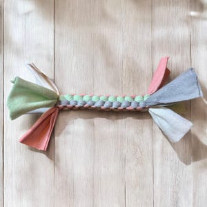 Könnte beinhalten: Ein geflochtenes Hundespielzeug mit mehreren Stoffstreifen in Grün-, Rosa- und Grautönen. Das Spielzeug ist aus weichem, strapazierfähigem Material gefertigt und perfekt zum Kauen und Apportieren.
