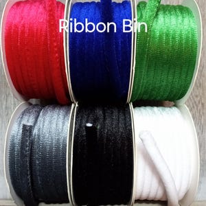 May include: Six spools of ribbon in various colours, including red, blue, green, grey, black, and white. The spools are arranged in a row on a wooden surface. The text "Ribbon Bin" is visible in the centre of the image.
