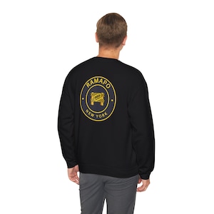 Op de afbeelding: Zwart sweatshirt met een geel en marineblauw cirkelvormig ontwerp op de rug. Het ontwerp toont "RAMAPO NEW YORK" langs de rand, met een wegversperring ontwerp en het woord "UNDER" in het midden.