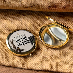 May include: A round gold-toned metal compact mirror with a hinged lid. The top features a graphic with the text "LET US DO THE TALKING," an illustration of Lady Justice holding scales, and a wooden gavel next to a stack of law books.