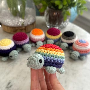 May include: A collection of handmade crochet turtles in various colors, including a rainbow turtle held in a hand. The turtles have small, black eyes and are displayed on a light-colored surface. The turtles are a craft item.