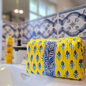 May include: A yellow quilted cosmetic bag with a blue and white floral pattern. The bag has a central blue panel with a leaf design. The bag is sitting on a white surface, with a blurred background of a bathroom.