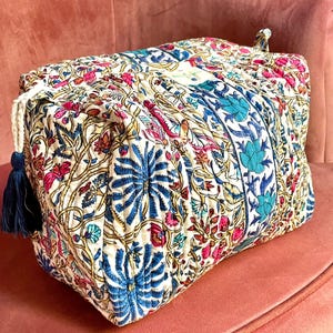 Peut inclure: Un sac matelassé rectangulaire avec un motif floral crème, bleu, rose et jaune. Le sac a une fermeture à cordon avec un pompon bleu marine. Le sac est posé sur une surface rose.