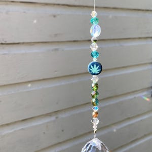 May include: A hanging crystal suncatcher with a variety of beads in clear, green, and blue hues. A dark blue disc with a white cannabis leaf design is featured. The suncatcher ends with a large, clear crystal ball.