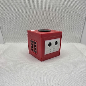 May include: A red and white cube-shaped object with a black circular top and a grid-like design on one side. The front features a white face-like design with two black circles and a horizontal line.