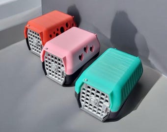 May include: Three small pet carriers in different colors: orange, pink, and teal. The carriers have a grid pattern on the front and are made of plastic.