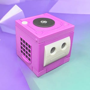 May include: A magenta-colored video game console with a circular disc tray and a white panel with two circular openings. The console has a square shape with a vent on the side and is set against a gradient background.