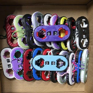May include: A collection of colorful combination lock toys in a cardboard box. The toys feature a variety of colors, including red, purple, blue, and green. Each lock has a unique number combination displayed. Some locks have a chain attached.