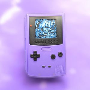 May include: A purple handheld gaming device with a screen displaying a blue cartoon character in a forest setting. The device has black buttons and the text "GAMES BOOK".