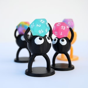May include: Three black 3D printed figurines with white eyes, each holding a different colored dice. The dice are a teal blue, pink, and rainbow color. The figurines are standing on black bases.
