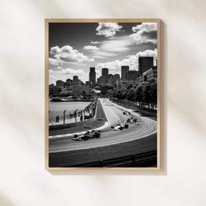 May include: Black and white framed print of a Formula 1 race in progress. The image captures race cars speeding along a track with a city skyline in the background. The sky is filled with clouds.