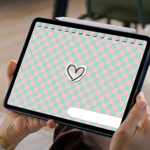 Puede incluir: Una tableta digital que muestra un diseño de cuaderno con un patrón de cuadros rosas y verdes. Una pegatina en forma de corazón está centrada en la pantalla. Un lápiz óptico descansa en la parte superior. La tableta la sostiene una persona.