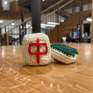 May include: Two crocheted mahjong tiles, one white with a red Chinese character, the other green and white.
