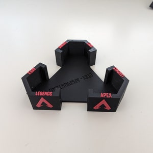 May include: A black, triangular game controller stand with three hexagonal holders. Each holder has the word "APEX" and "LEGENDS" in red, along with a red triangle logo. The stand is made of plastic.