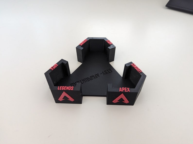 May include: A black, triangular game controller stand with three hexagonal holders. Each holder has the word "APEX" and "LEGENDS" in red, along with a red triangle logo. The stand is made of plastic.