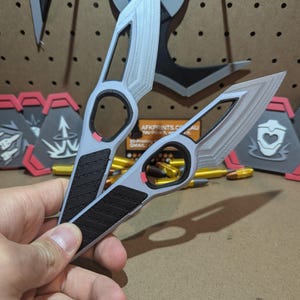 May include: Two gray 3D printed throwing knives with black and red accents. The knives have a unique design with a hole in the handle. The knives are held in a hand.